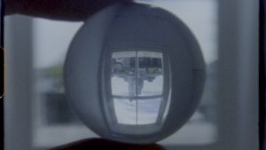 Still from 'Lucida' by Suki Chan. © Suki Chan. A small ball shaped object held between fingers through which an upside -down building can be viewed through a window.