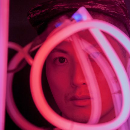 Photograph of artist Samson Young looking through a circular pink neon light.