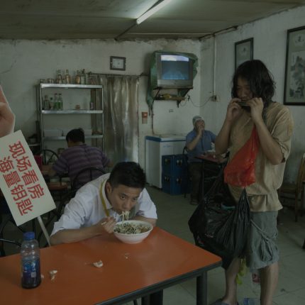 Man sat at table eating noodles with placard to the left of him. Next to the table a man dress in t-shirt and shorts is playing a harmonica.