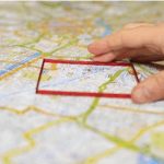 A hand holding an outline of a red square, which is being held on a map.