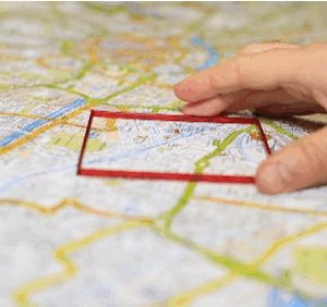A hand holding an outline of a red square, which is being held on a map.
