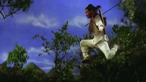 Video still of a martial arts man in the air about to land on the ground. There are trees in the background.