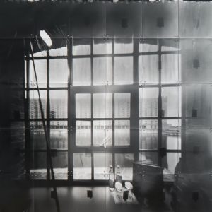 Black and white photograph of a window in the foreground and the view of industrial buildings in outside of the window.