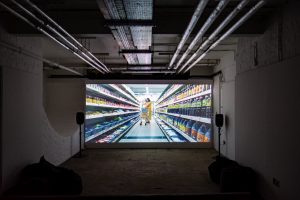 A video installation showing a woman in a supermarket. The screen is in a basement with speakers at either side and exposed pipes on the ceiling.