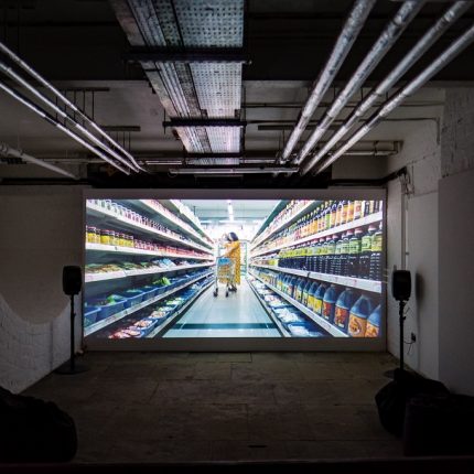 A video installation showing a woman in a supermarket. The screen is in a basement with speakers at either side and exposed pipes on the ceiling.