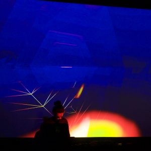 Man stood in front of a large screen which is mainly blue with a semicircle of yellow light at the bottom of the screen with a red halo around the semicircle.