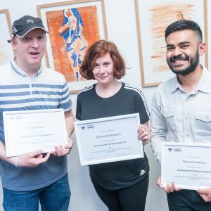 Three people holding framed awards, stood in front of paintings.