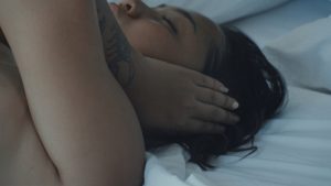 Video still a woman lying down with her hands over her ears.