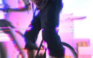 Pink distorted background, in the foreground there is a close-up of someone on a bike. Upper body of the person is not shown