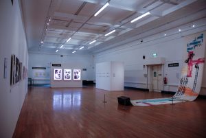 Gallery photograph. In the distance are 3 lightboxes of 3 people's faces. On the right is an artwork on material which is draped on the floor.