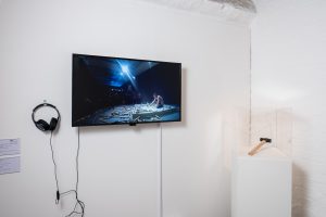 TV screen on white wall showing a man sat on a platform surrounded by hammers. To right of the screen a hammer on a plinth under a perspex case.