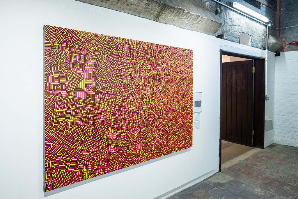a photograph of a large painting hung on a white wall. The painting shows a detailed, abstract map, in yellow, red and black.