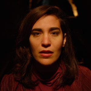 Head and shoulders shot of a woman staring into the camera. The background behind her is black except for a small line of light in the distance.