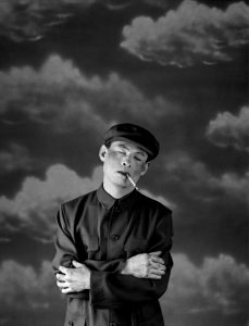 Black and white photograph. A man dress in a uniform with a cigarette hanging from the side of his mouth, stands with cross arms across his body and his eyes shut. The background depicts images of clouds.