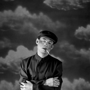 Black and white photograph. A man dress in a uniform with a cigarette hanging from the side of his mouth, stands with cross arms across his body and his eyes shut. The background depicts images of clouds.