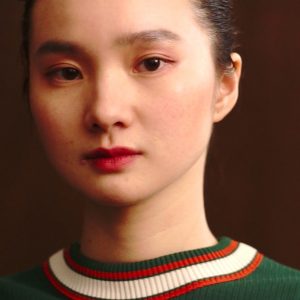 A still image from a film. The image shows a head and shoulders shot of a woman facing the camera. The woman has a green jumper on and her hair tied back.