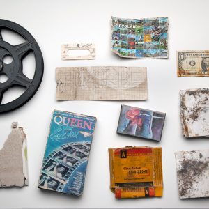 Photograph of what appears to be disregarded objects, including an American Dollar, a film reel, a photographic slide and a Queen concert ticket.