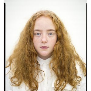 Head and shoulders shot of a girl. The girl is staring straight at the camera, she has long curly auburn hair. The girl is in a white shirt and is against a white background.
