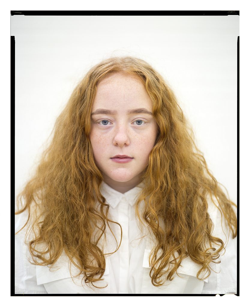 Head and shoulders shot of a girl. The girl is staring straight at the camera, she has long curly auburn hair. The girl is in a white shirt and is against a white background.