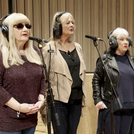 A film still of three women stood singing at microphones.