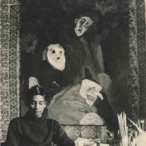 A black and white photograph of African-Indian artist Albert Adams, likely in his 20s at the time, sat in front of a large semi abstract, semi figurative painting in the studio.