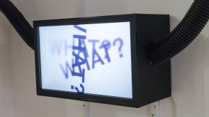 A photograph of a television facing forwards and slightly to the left. The television is a black box shape and mounted to a white wall. Coming out from the left and right side of the television are two black tubes. On the screen, the fading words ‘WHAT?’ are repeated three times overlapping on top of each other at different angles.