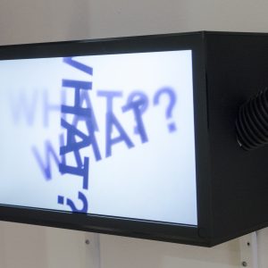 A photograph of a television facing forwards and slightly to the left. The television is a black box shape and mounted to a white wall. Coming out from the left and right side of the television are two black tubes. On the screen, the fading words ‘WHAT?’ are repeated three times overlapping on top of each other at different angles.
