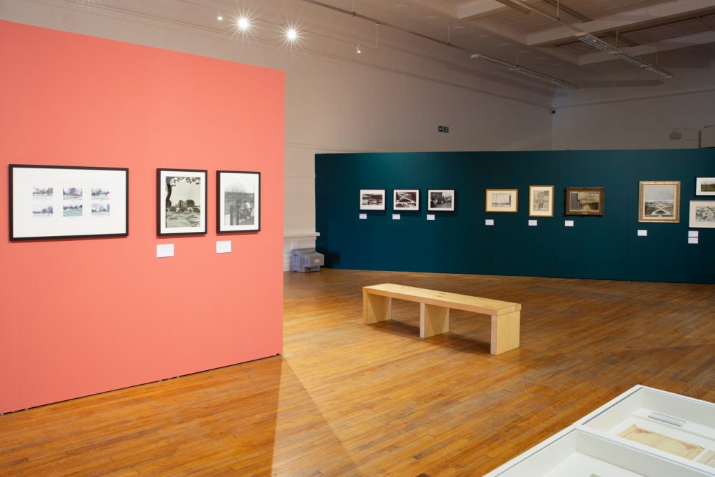 Interior photo of a gallery exhibition. The image shows a pink wall and a green wall, with small framed artworks hung on them.
