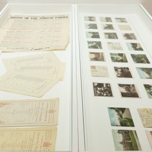 Two long, white, flat, glass topped display cases. In the left case are archive materials - posters and documents from music performances in Peel Park. The paper is yellowed and aged. In the right, a number of postcards, both face up and face down, showing images and written inscriptions. The postcards are of Peel Park and Salford, over the last 100 years.