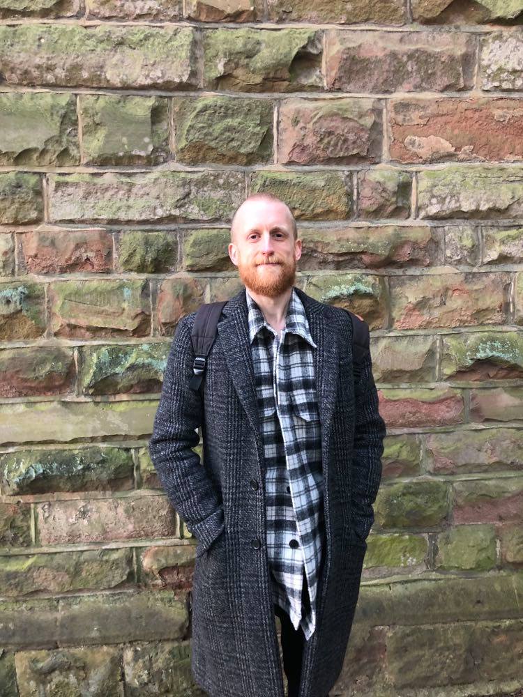 A photograph of Greg Salter, from the waist up against a brick background. 