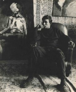 a young man sits in a large armchair in front of a painting. He faces the camera with a thoughtful expression. The photo is in sepia black and white