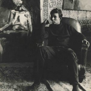 a young man sits in a large armchair in front of a painting. He faces the camera with a thoughtful expression. The photo is in sepia black and white