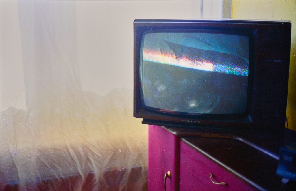 A box style television sits on the corner of a bright pink cabinet. The screen shows a grainy image. The bottom half is black, in the top half a pair of eyes and hat are just visible. Some reflections of the room can be seen. Behind, a thin white fabric covers a window, allowing a soft white and yellow glow of light into the room.