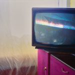 A box style television sits on the corner of a bright pink cabinet. The screen shows a grainy image. The bottom half is black, in the top half a pair of eyes and hat are just visible. Some reflections of the room can be seen. Behind, a thin white fabric covers a window, allowing a soft white and yellow glow of light into the room.