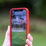 a hand holds up a smartphone, on the screen a digital image of a bandstand is superimposed on the background of a park