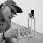 A black and white image shows the artist looking over small figure like statues on small woden plint. The artist is holding the object on the furthest left, as if to move it.