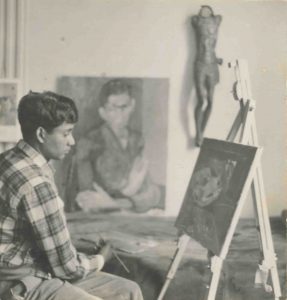 A black and white photograph shows a young Albert Adams, in profile, sat holding a painbrush and facing a small easel with a canvas.