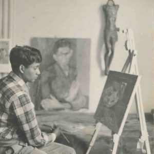 A black and white photograph shows a young Albert Adams, in profile, sat holding a painbrush and facing a small easel with a canvas.