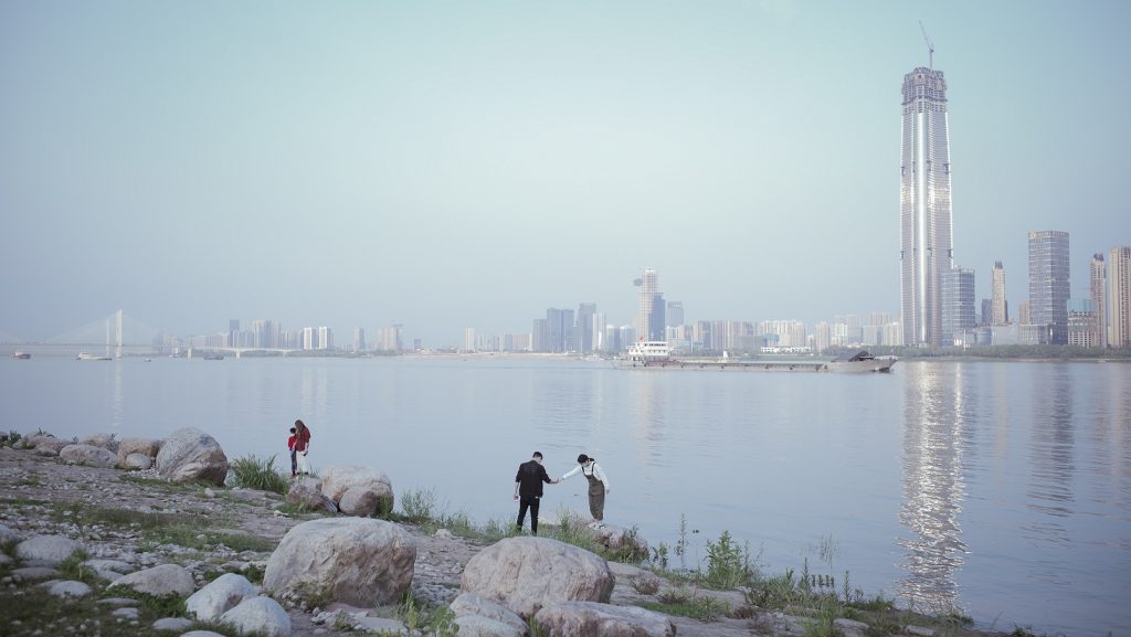 An images shows a man and a woman reaching out for each other and holding hands by the water front. Behind them the Wuhan skyline rises into the blue sky.