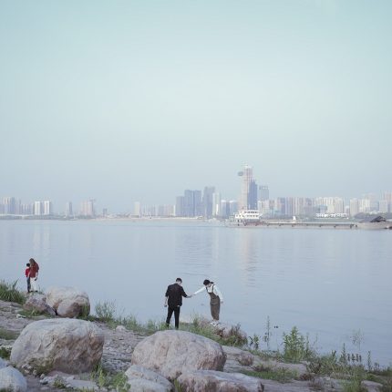 An images shows a man and a woman reaching out for each other and holding hands by the water front. Behind them the Wuhan skyline rises into the blue sky.