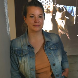 Portrait of Emily Speed, shown from the waist up. She is a white woman with light hair, wearing an orange shirt and light wash denim jacket.