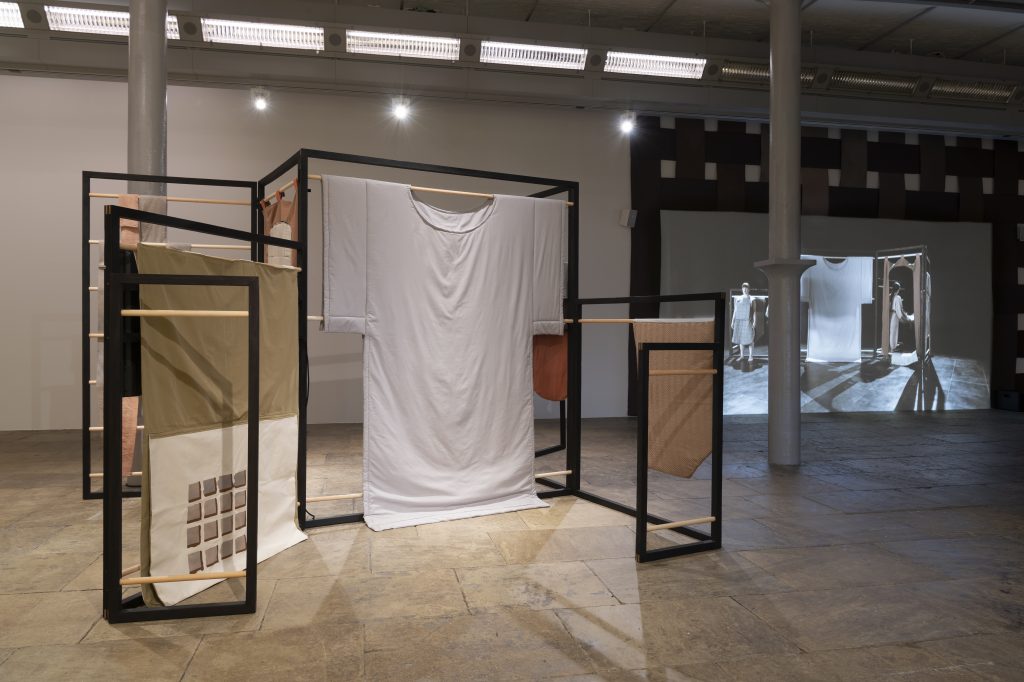Image of 'Flatland' by Emily Speed, installed at Tate Liverpool. The image shows a white tshirt recreated at person height, suspended amongst other fabrics on a black frame. 