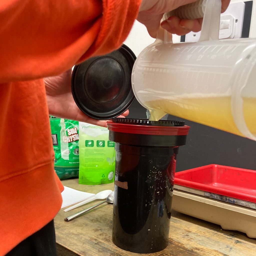 A person in an orange jumper pours plant-based developing solution into a film developing tank.