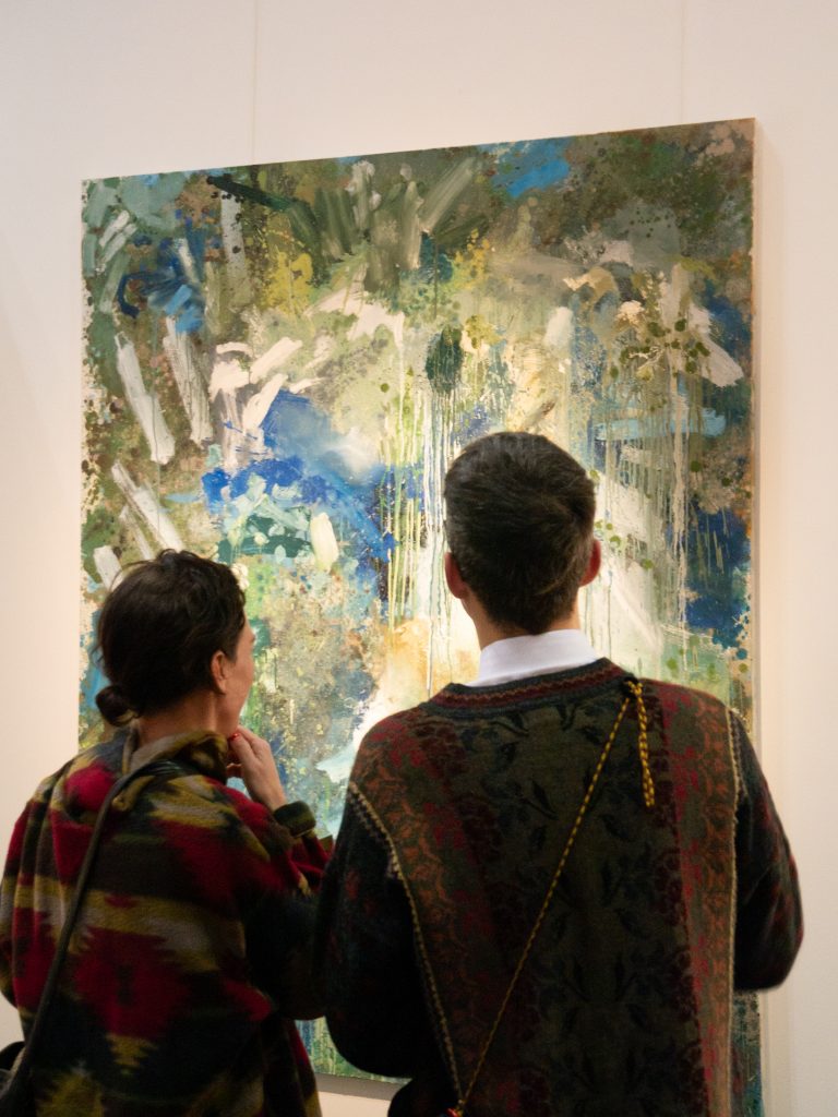 The head and shoulders of two people, from the back, stood next to each other. They are admiring a painting on the wall. The painting is abstract greens and blues.