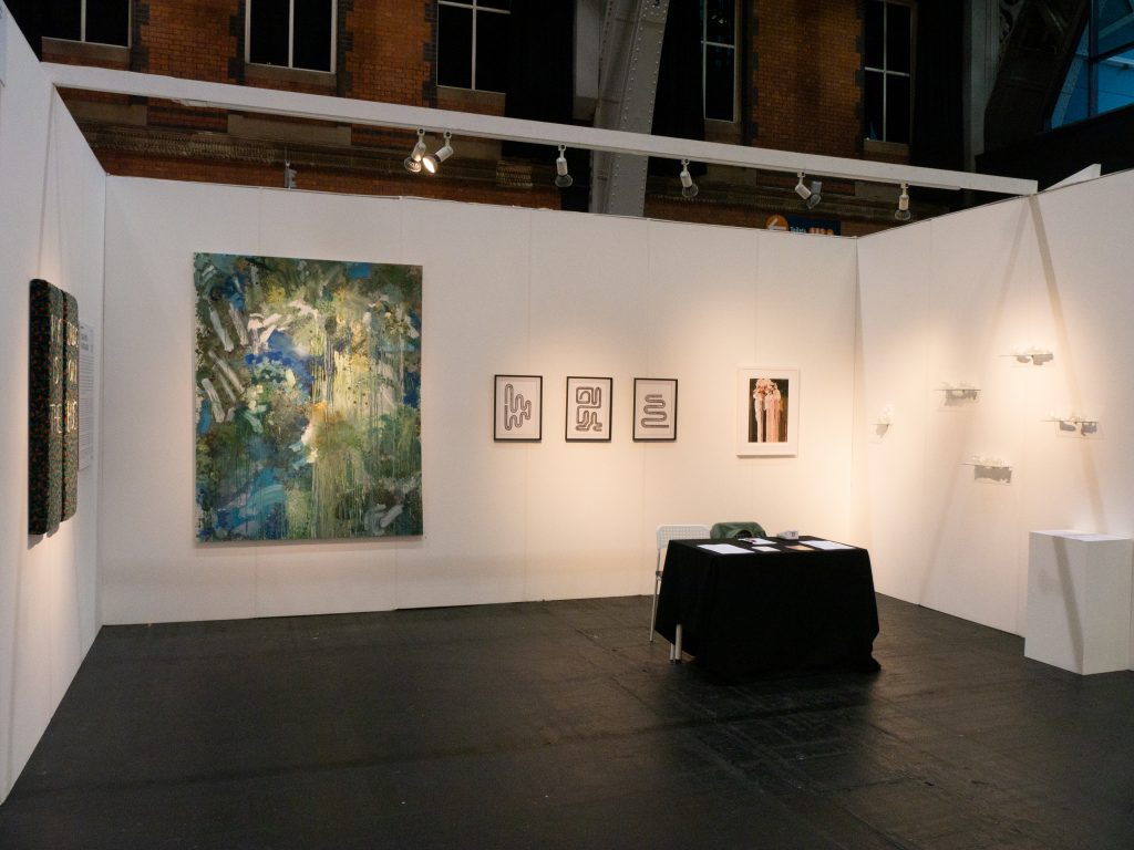 A gallery booth at the manchester contemporary art fair 2023. A dark grey black floor and three white walls, with a small chair and table at the centre. On the walls are various artworks, on canvas, in frames, and mini sculptures on shelves.