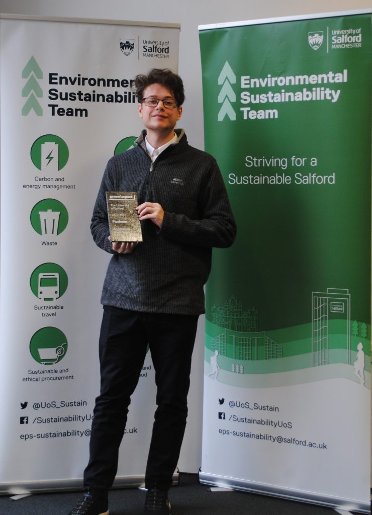 Team Assistant Sam Parker holding the Green Impact award in front of two University sustaininability banners.