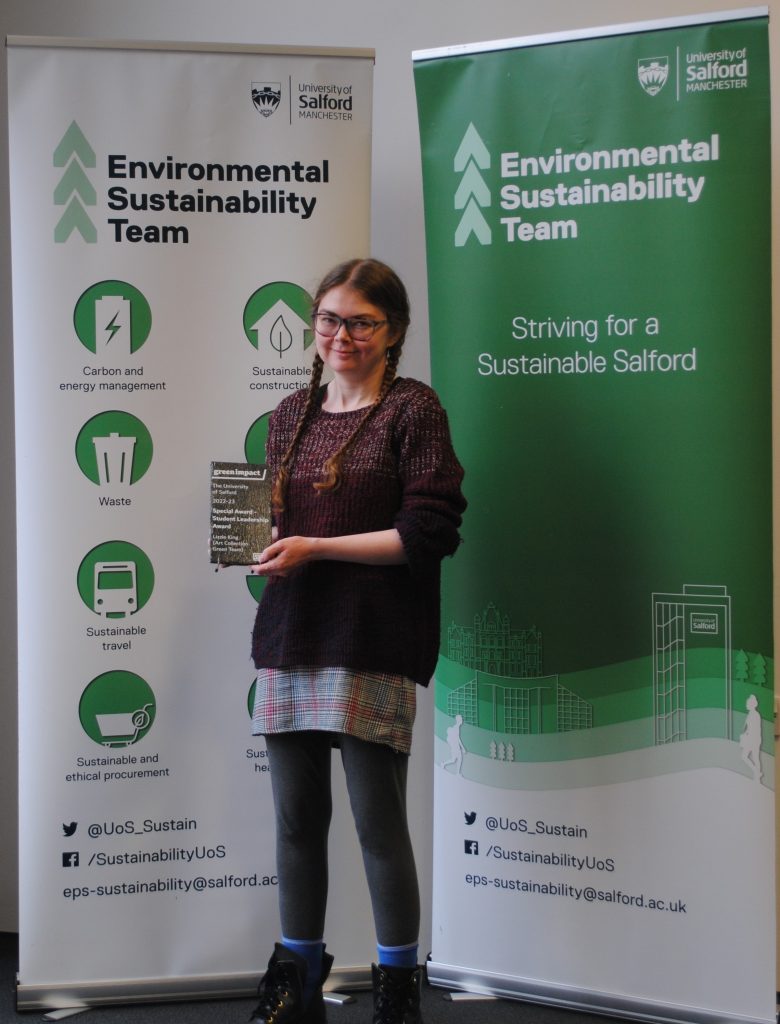 Lizzie King holds her Green Impact award, stood in front of two sustainability banners.