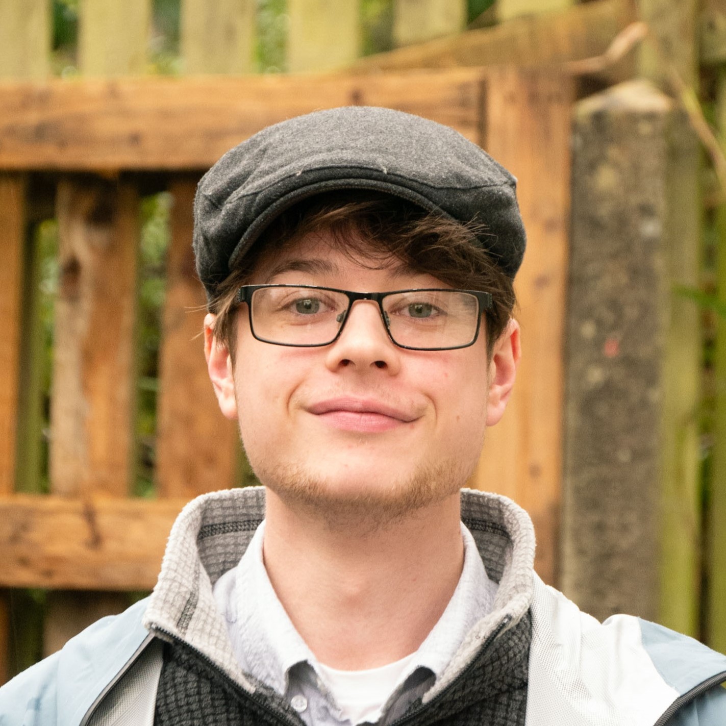 A picture of our newest team assistant Sam Parker. Wearing a grey flat clap, glasses, shirt, jumper, and coat. He is outside, with a rustic wooden fence connected to a stone pillar behind him.