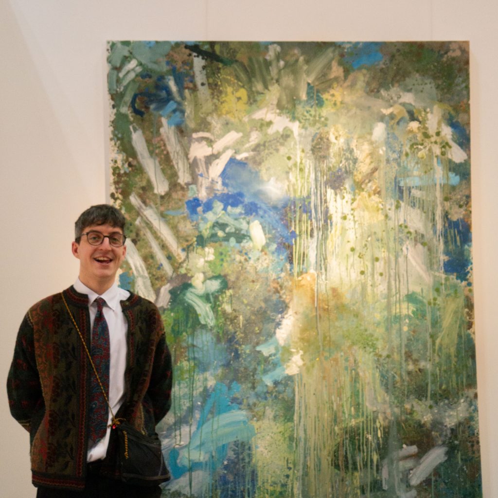 Photograph of Graduate Scholar Adam Rawlinson at the Manchester Contemporary 2023, stood in front of his oil painting 'It's Nice To Be Alive' (2023). Photograph taken by Sam Parker.