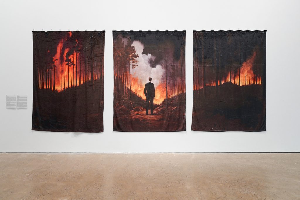 Mishka Henner's large scale work Executivie Decision, comprised of three large blankets hung on a white wall. The blankets show a forst on fire, with a man in a buisness suit looking out towards the destruction in the style of Caspar David Friedrich's Wanderer Above The Sea of Fog.